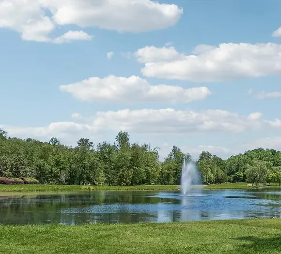 Landscaped pond with a white spray fountain, a line of hardwoods and pines behind it and a strip of mown lawn in front, under a partly cloudy sky