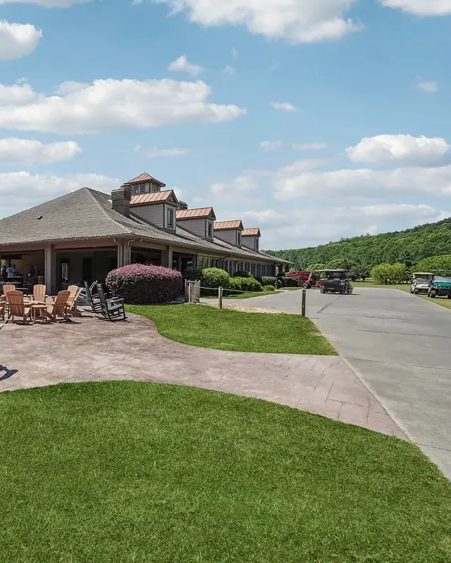 Long clubhouse with a shingle roof and copper-capped dormers, a stamped-concrete patio with Adirondack chairs, and a wide mown lawn below wooded hills