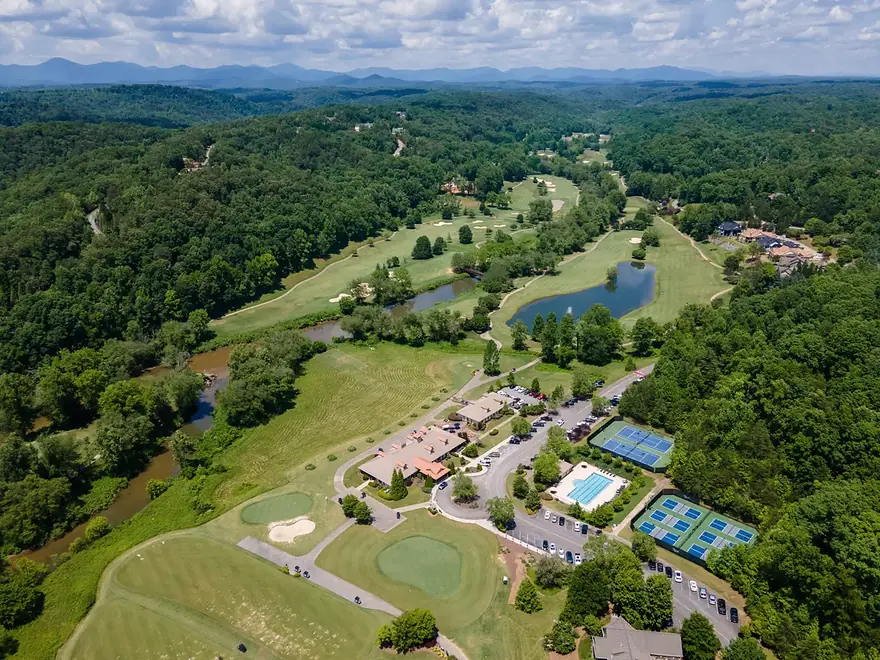 Aerial view over golf fairways, ponds and a river to forested hills and blue ridges, with a clubhouse, pool and tennis courts in the foreground