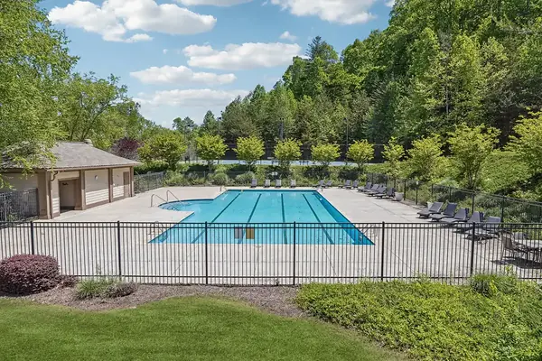 Fenced community pool with lap lanes and lounge chairs, a pool house at left and tennis courts and wooded hills behind