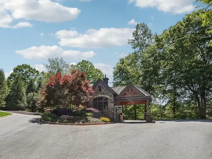 Stone gatehouse with a timber porte-cochere, red-leaf shrubs in a planted median and a wide paved entry drive