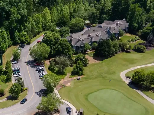 Aerial view of a cluster of shingle-roofed homes among hardwoods above a golf green, a curving cart path and a parking area
