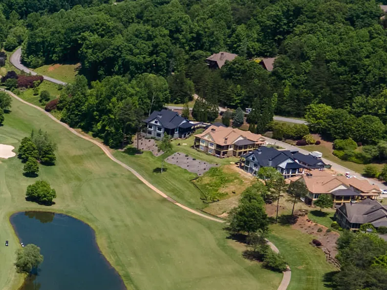 Aerial view of large homes with dark and terracotta roofs set along a fairway, with a cart path, a pond and hardwoods