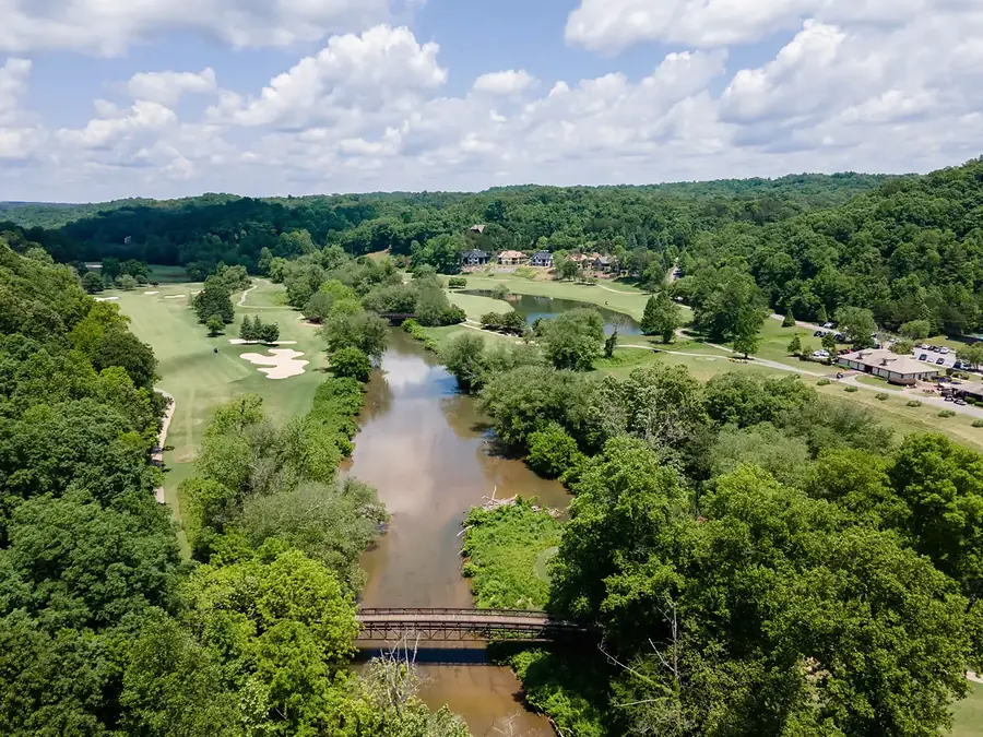 Aerial view of a river running between wooded banks, a footbridge in the foreground and golf fairways with sand bunkers on one bank